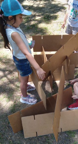 Una nena construyendo una estructura triangular de cartón al aire libre